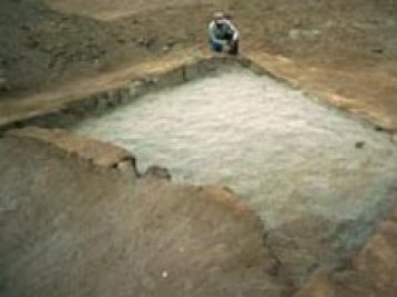 A rectangular excavated pit with an archaeologist sitting in the corner.